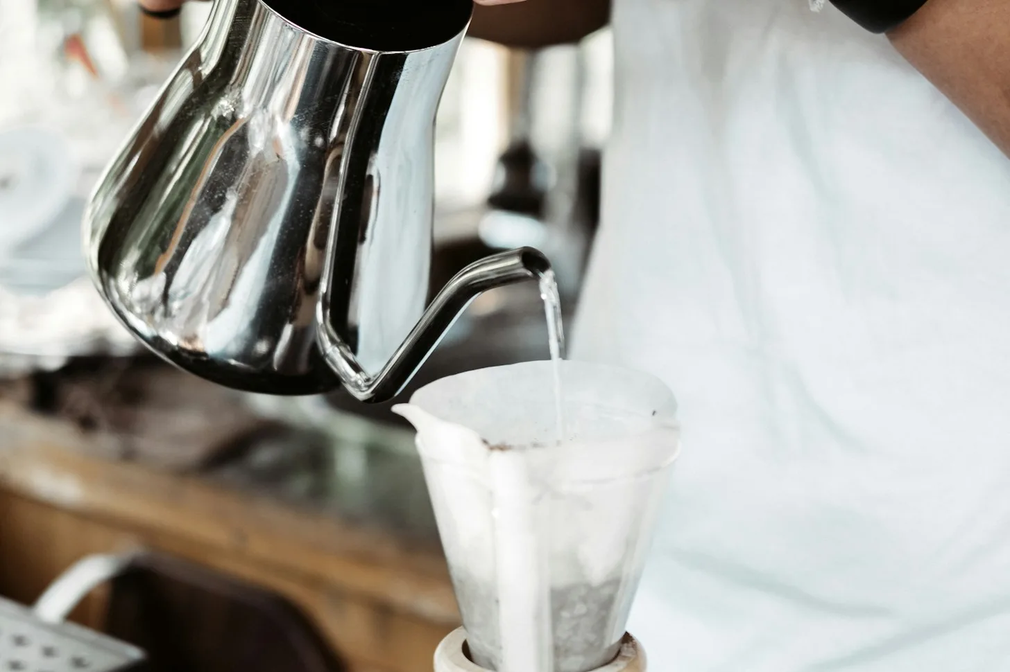 Hands pouring hot water through a paper filter into a glass coffee brewer